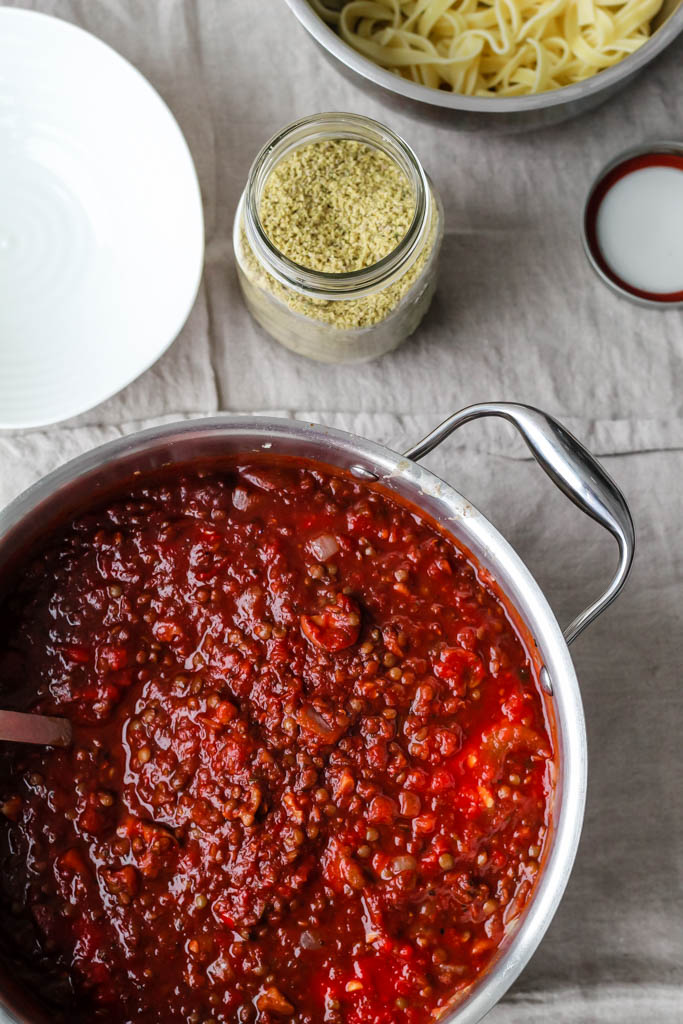 An image showing a pot full of lentil mushroom bolognese pasta sauce surrounded by pasta and vegan parmesan. 