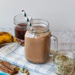 straight on shot of the dirty chai smoothie in a glass surrounded by a jar of hemp, coffee, ripe banana, and cinnamon sticks.