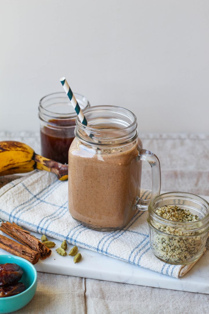 straight on shot of the dirty chai smoothie in a glass surrounded by a jar of hemp, coffee, ripe banana, and cinnamon sticks.