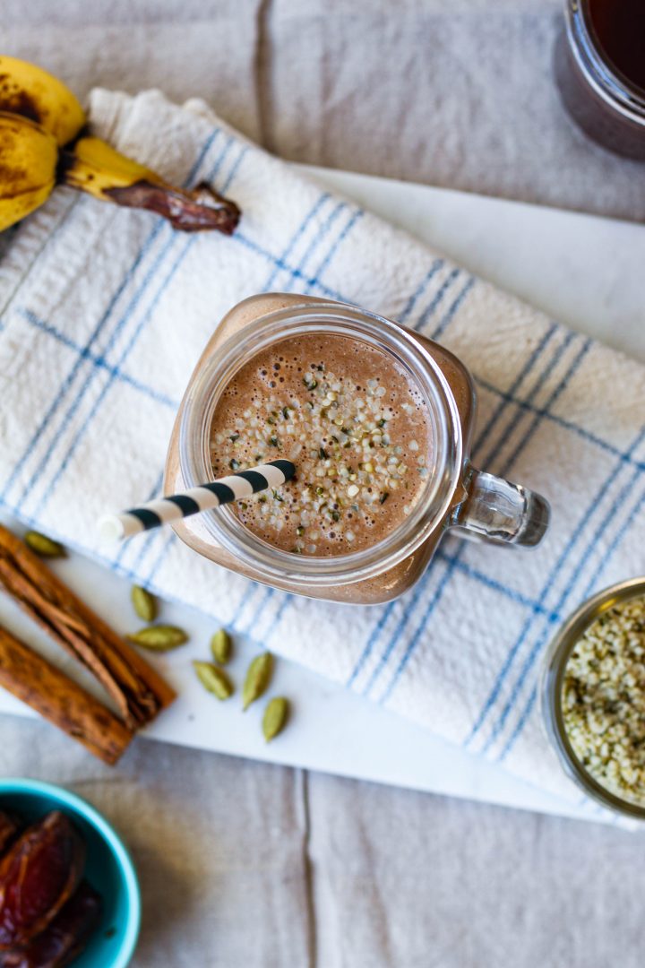 overhead shot of the dirty chai smoothie on a marble board with spices, hemp, and banana surrounding 