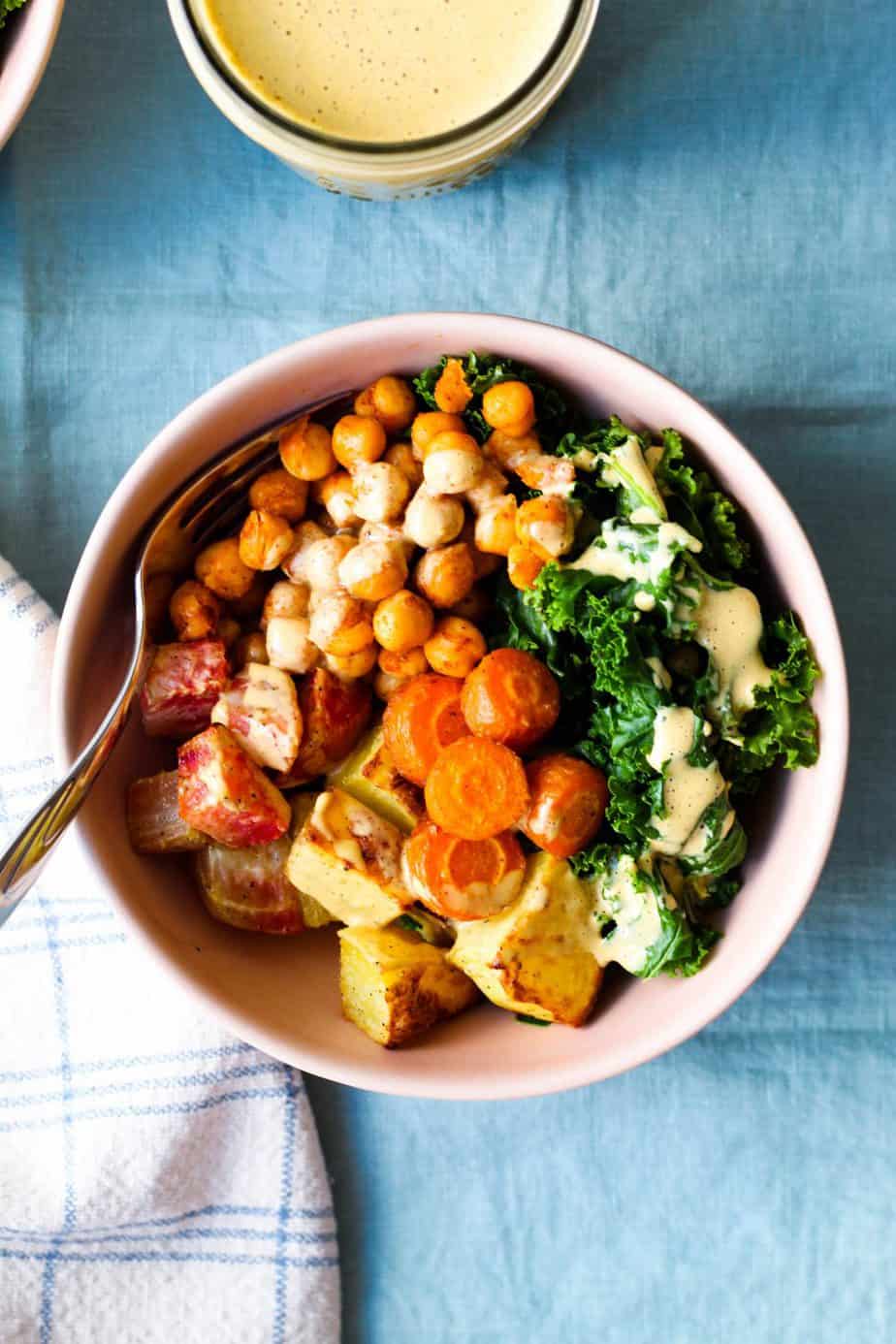 overhead shot of a bowl with roasted potato, beets, carrots, and steamed kale, roasted chickpeas, and nutritional yeast dressing drizzled over top