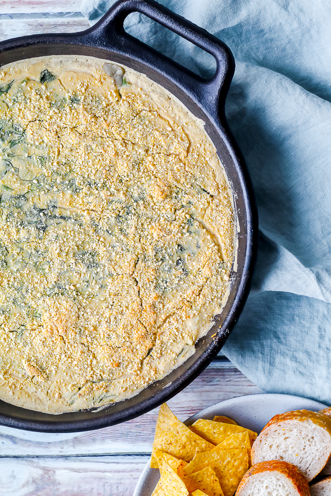 overhead shot of the fresh out of the oven white bean spinach artichoke dip in a cast iron skillet with baguette and chips surrounding it 