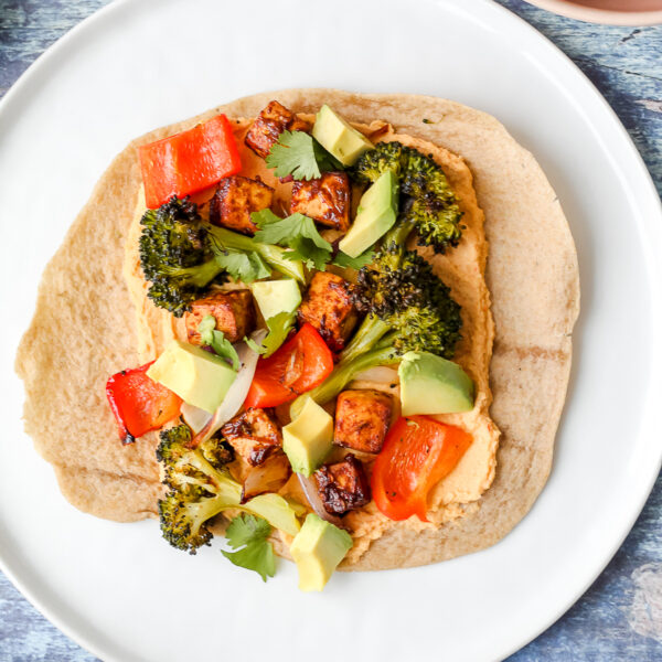 an overhead shot of a tortilla spread with chipotle hummus and topped with the roasted vegetables, chili lime tofu, and avocado