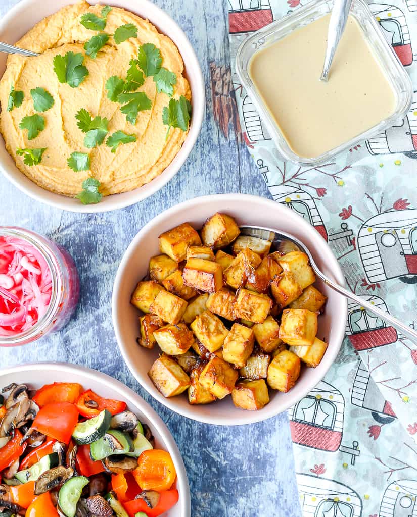 an overhead shot of bowls of red curry peanut hummus, tofu, roasted veggies, pickled onions, and pita sauce on a blue backdrop 