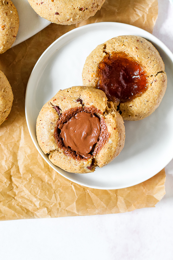 photo of one chocolate hazelnut thumbprint cookie and one jam thumbprint cookie on a white plate 