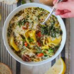 an overhead photo of a bowl of creamy chicken pesto tortellini soup surrounded by lemon wedges and sourdough buns with butter