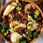 overhead shot of the fall harvest beet, apple, pomegranate, and avocado kale salad in a wooden bowl on a grey backdrop