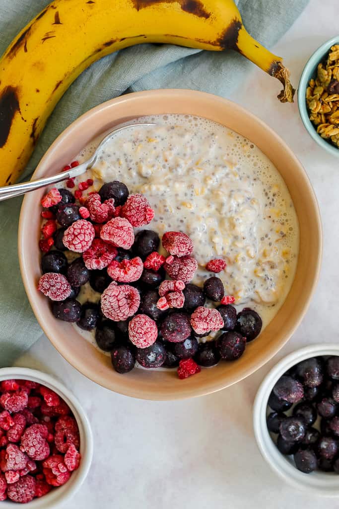 overhead shot of the overnight oats surrounded by a banana, a small dish of granola, and small dishes of blueberries and raspberries