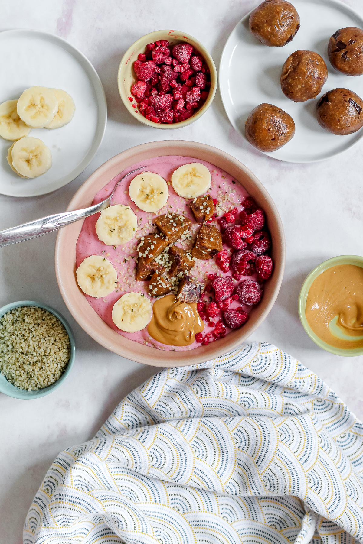 high angle overhead shot of the smoothie bowl surrounded by a plate of energy balls, raspberries, banana, cashew butter, and hemp on a light pink backdrop