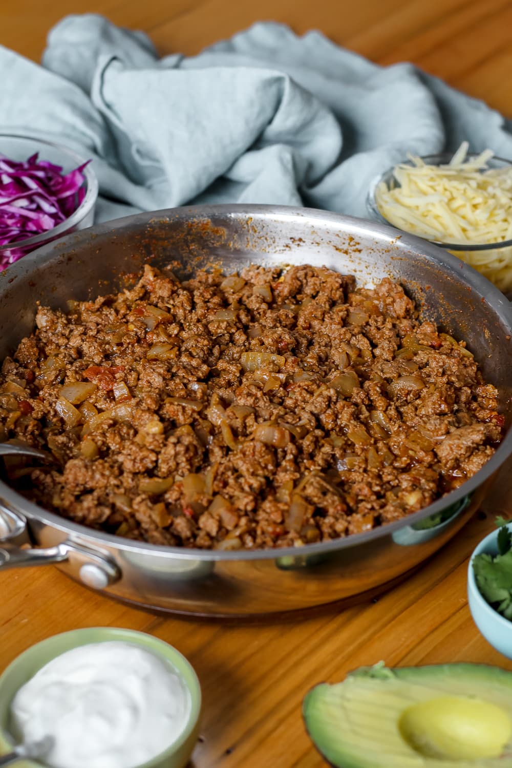 angled overhead shot of a skillet of beef taco meat surrounded by grated cheese, purple cabbage, avocado, cilantro, and sour cream.