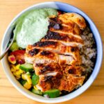 a close up view of a bowl of sliced chicken breast, brown rice, summer corn, cucumber, and tomato salad, and pesto Greek yogurt sauce
