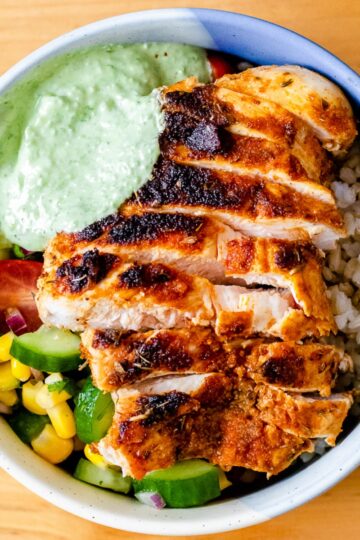 a close up view of a bowl of sliced chicken breast, brown rice, summer corn, cucumber, and tomato salad, and pesto Greek yogurt sauce