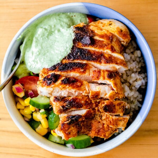 a close up view of a bowl of sliced chicken breast, brown rice, summer corn, cucumber, and tomato salad, and pesto Greek yogurt sauce
