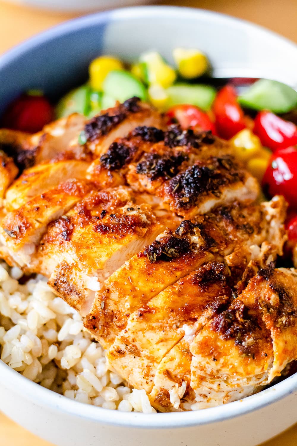 A close up photo of a sliced chicken breast in a bowl along with brown rice and tomato corn cucumber salad