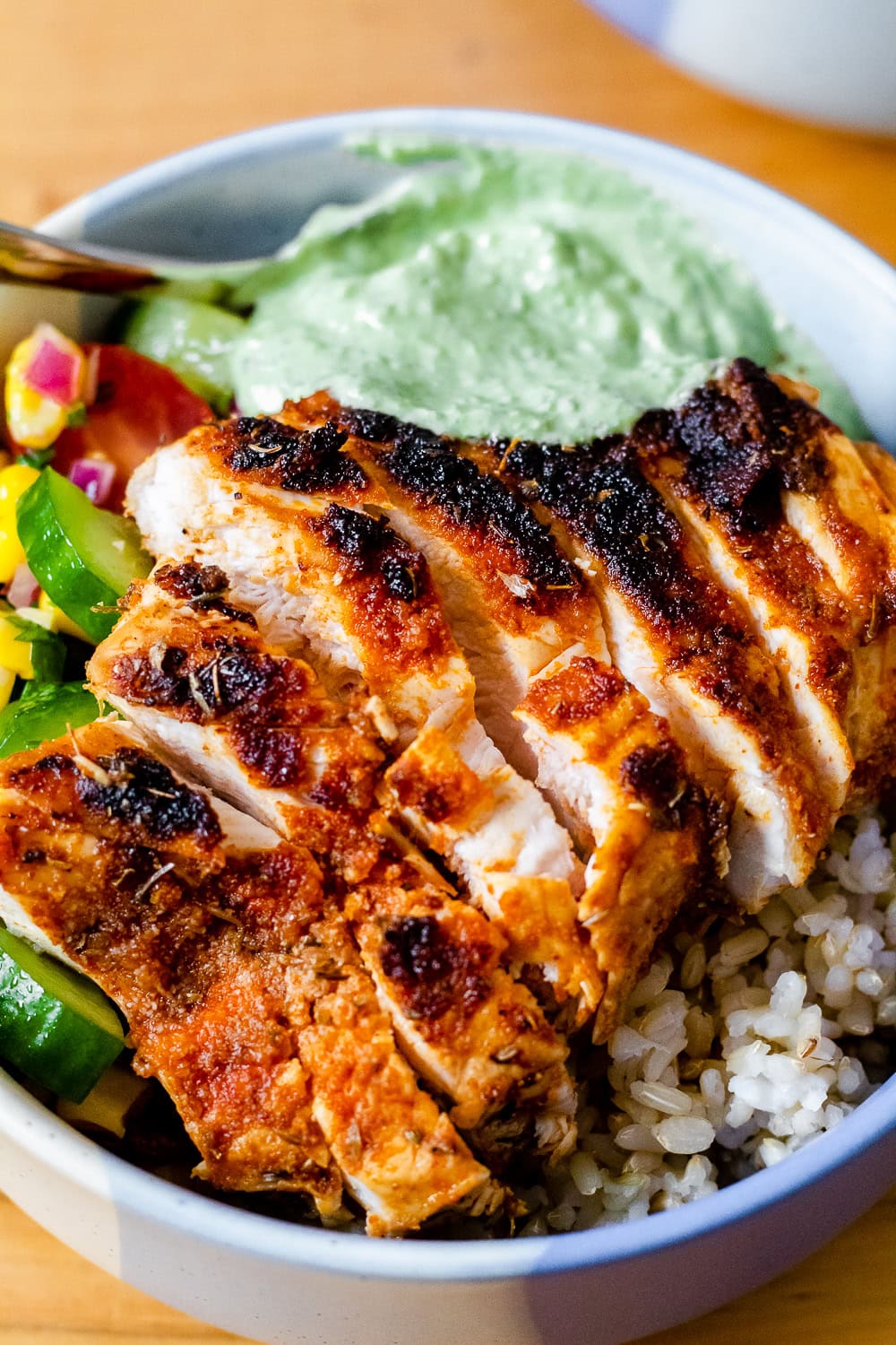 close up overhead view of sliced chicken breast, pesto sauce, summer tomato cucumber salad, and brown rice in a blue and white bowl