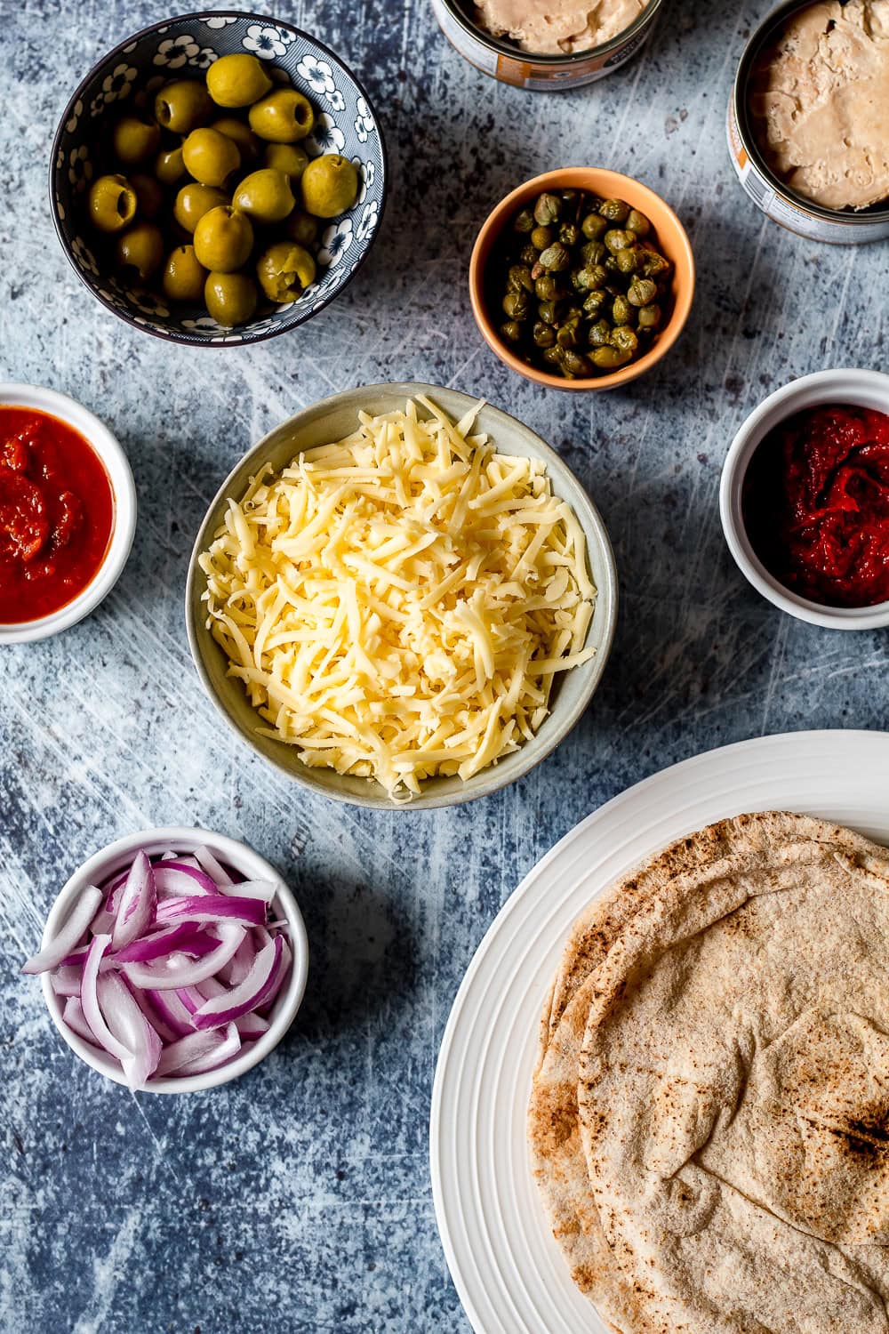 an overhead photo of all of the ingredients for tuna pizza, including pitas, cheese, red onion, canned tunoa, capers, green olives, and pizza sauce