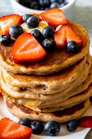 a close up photo of a stack of the pancakes on a white plate. The pancakes are topped with butter, maple syrup, strawberries, and blueberries