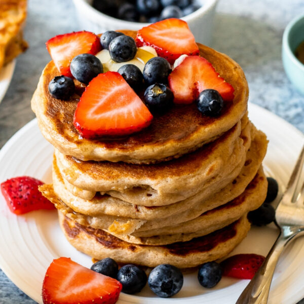 a close up photo of a stack of the pancakes on a white plate. The pancakes are topped with butter, maple syrup, strawberries, and blueberries